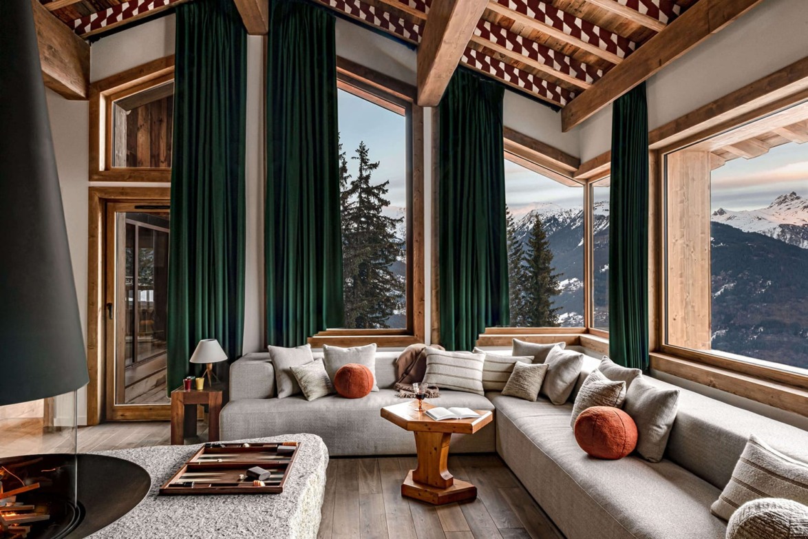 Cozy mountain chalet living room with large windows, green curtains, and modern furniture overlooking snowy landscape.