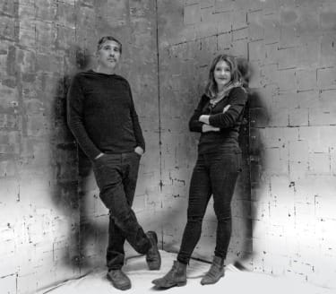 Two people standing against a textured wall, both wearing dark clothing, looking at the camera in a monochrome setting.