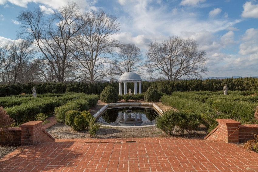 Garden with a brick path leading to a reflective pond and a small white pavilion in the background, surrounded by trees.