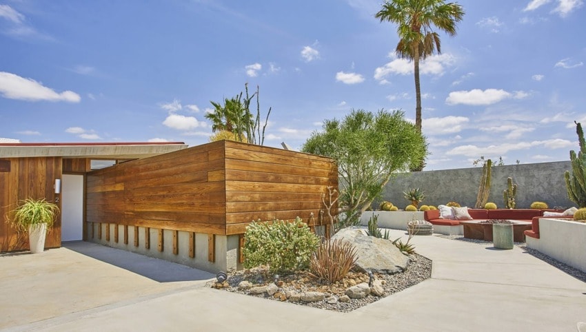 Modern house exterior with wooden paneling, desert landscaping, and outdoor seating under a blue sky with palm trees.