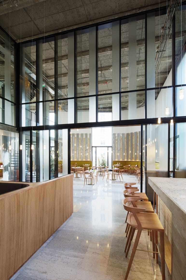 Modern cafe interior with large windows, wooden furnishings, and high ceilings, featuring a cozy seating area.