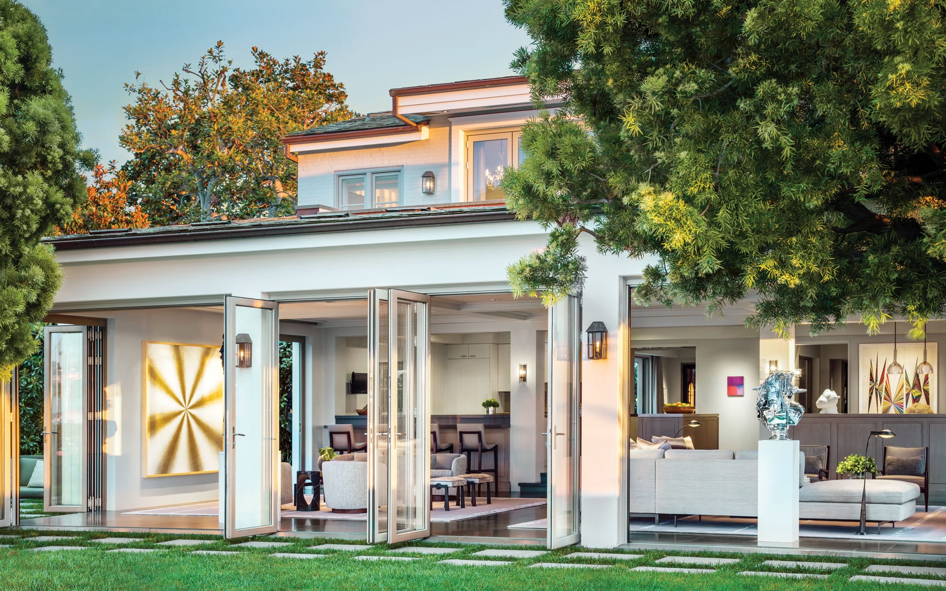 Modern house exterior with open glass doors leading to a stylish interior, surrounded by lush greenery and sunlight.