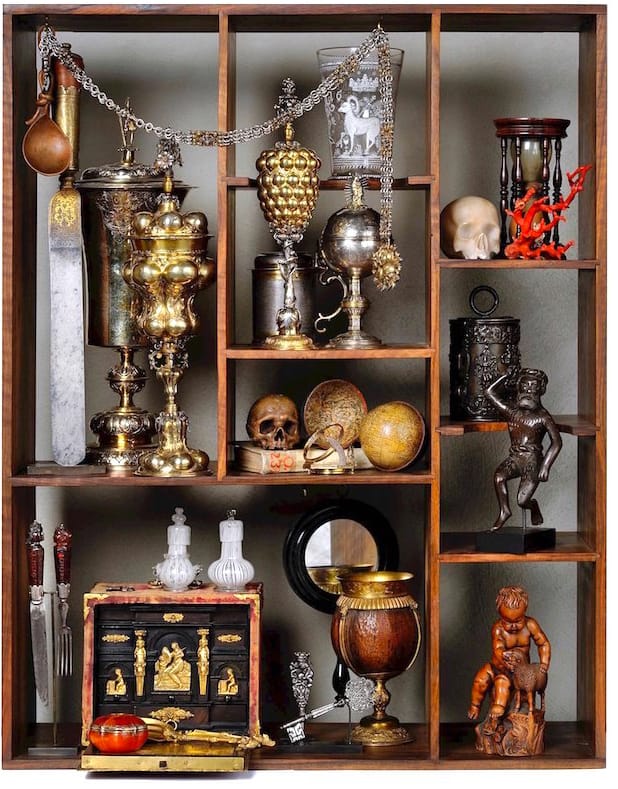 Collection of vintage and ornate objects including goblets, skulls, a monkey statue, and decorative boxes in a wooden shelf.