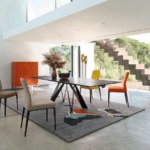 Modern dining room with a table, four chairs, abstract rug, and large windows overlooking greenery.