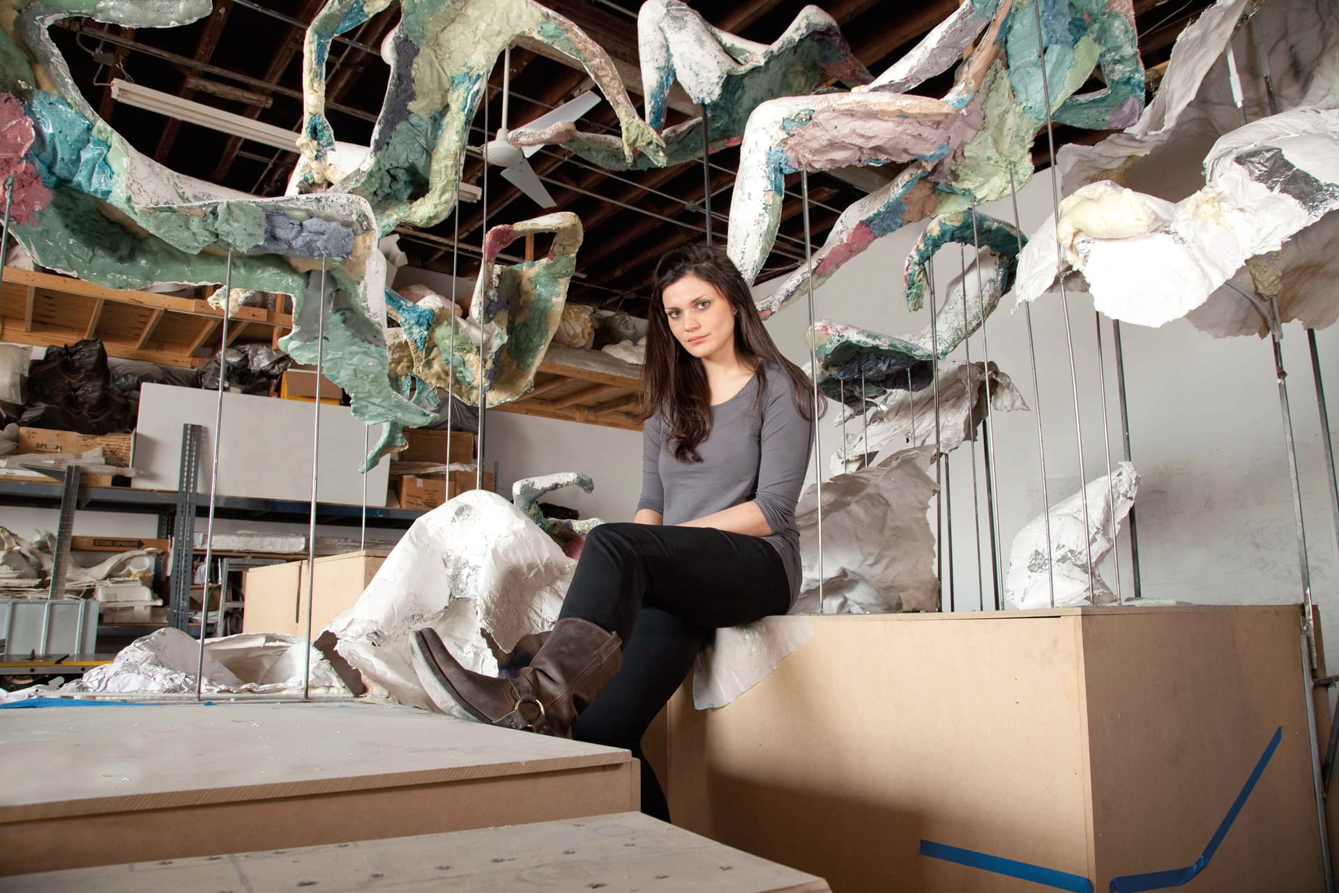 Woman seated in an art studio surrounded by abstract sculptures and colorful hanging paper art pieces.