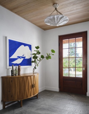 Modern foyer with wooden console, art on wall, plant vase, and wooden door with glass panels.