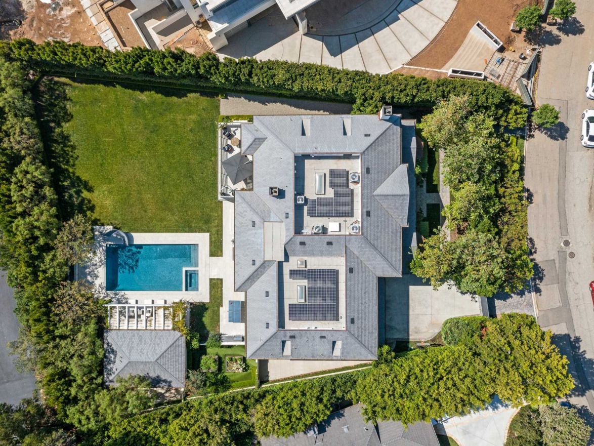 Aerial view of a large modern house with a pool and surrounding trees in a suburban neighborhood.