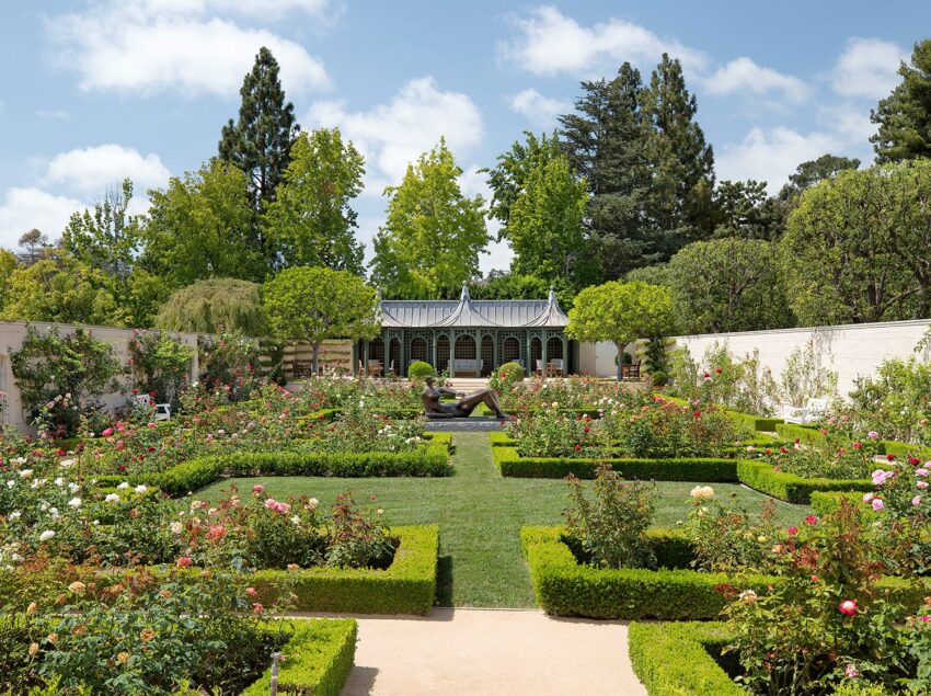 A formal garden with a symmetrical design, green hedges, colorful flowers, and a central statue under a blue sky.