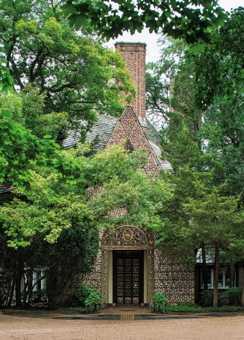 Charming brick house with ornate doorway surrounded by lush green trees and bushes on a gravel path.