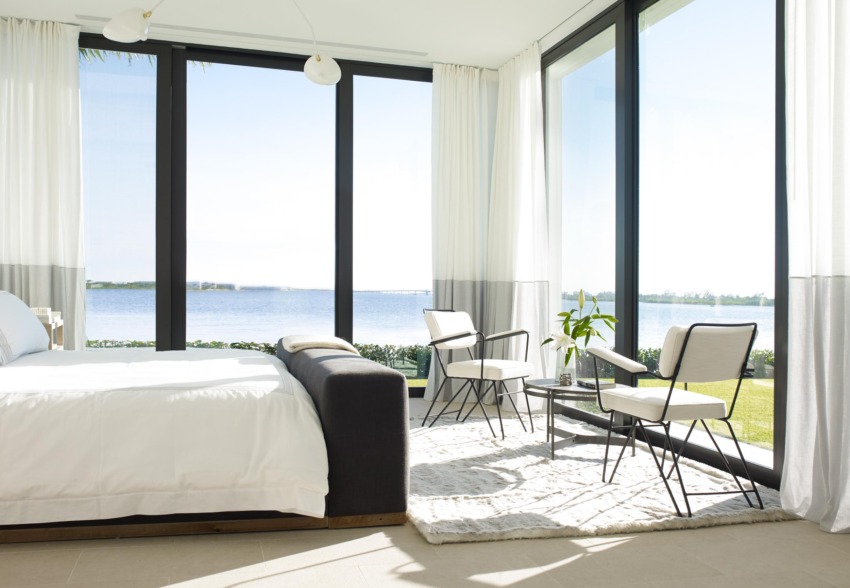 Modern bedroom with large windows overlooking a scenic lake view, featuring a cozy bed and elegant seating area with natural light.