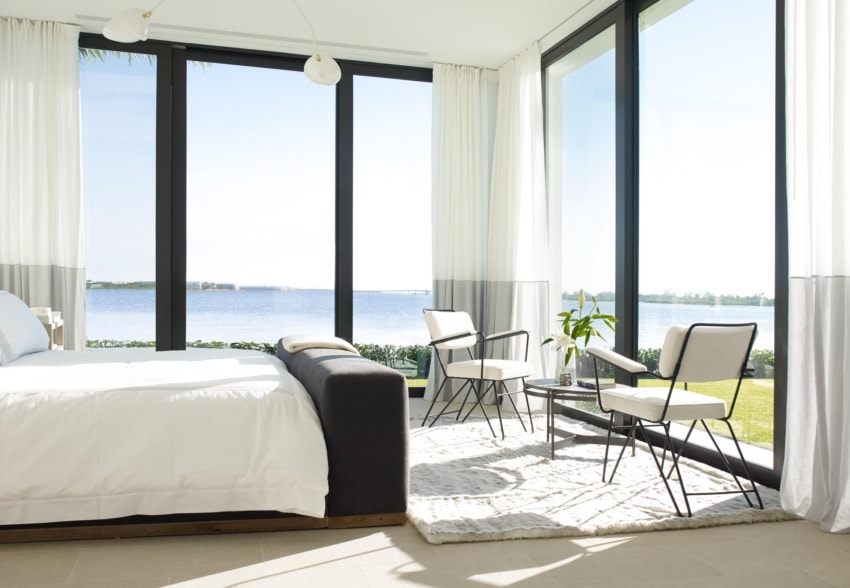 Modern bedroom with large windows overlooking a scenic lake view, featuring a cozy bed and elegant seating area with natural light.