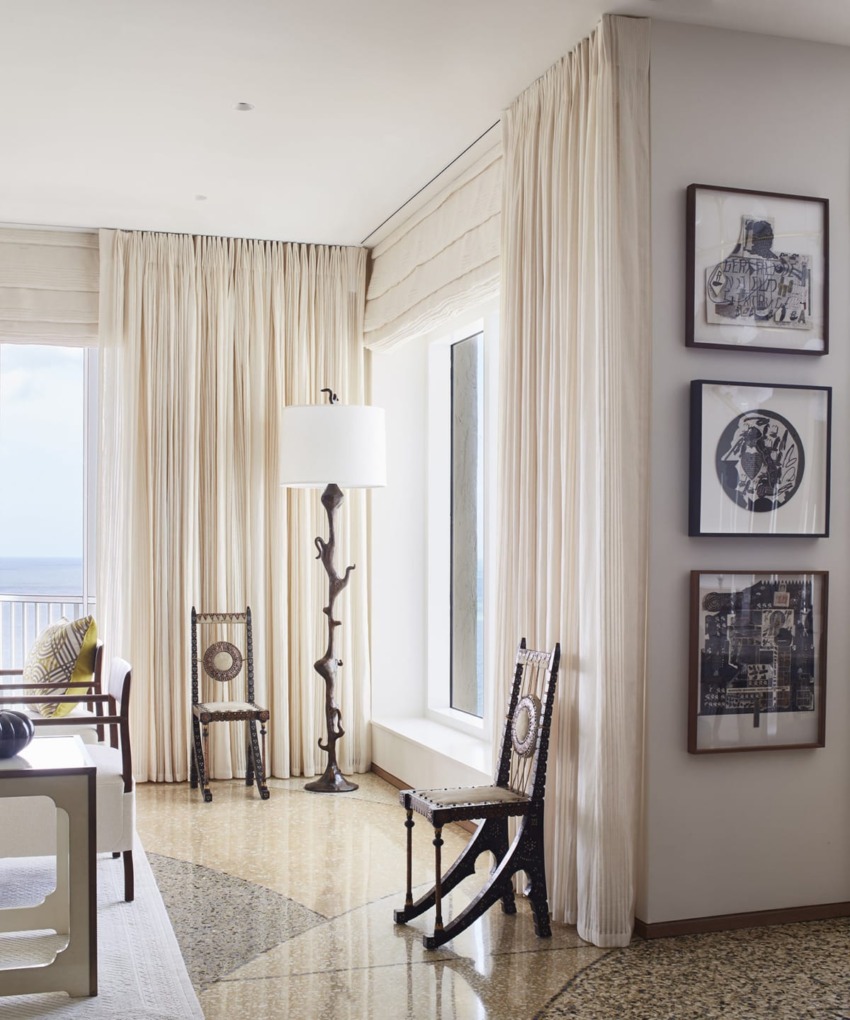 Bright living room with large windows, cream curtains, decorative chairs, a unique floor lamp, and framed artwork on the wall.