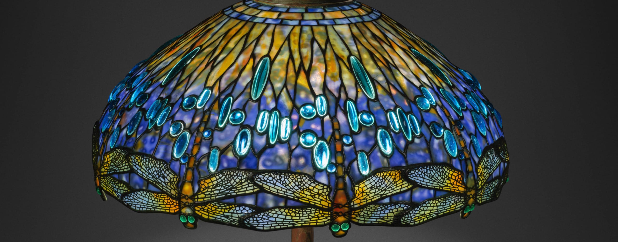 Stained glass lampshade with intricate dragonfly and gemstone pattern in blue and yellow tones on a gray background.