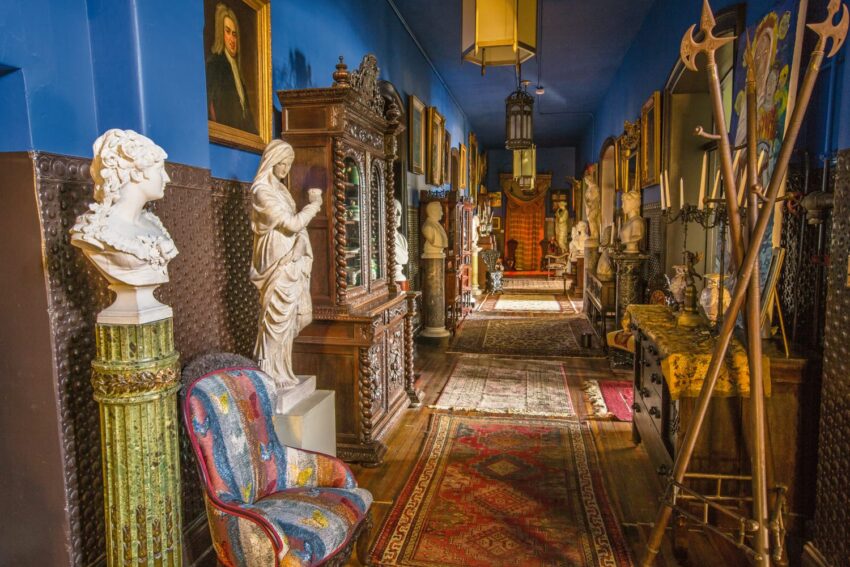 Eclectic hallway with statues, ornate furniture, colorful chair, and portraits on blue walls.