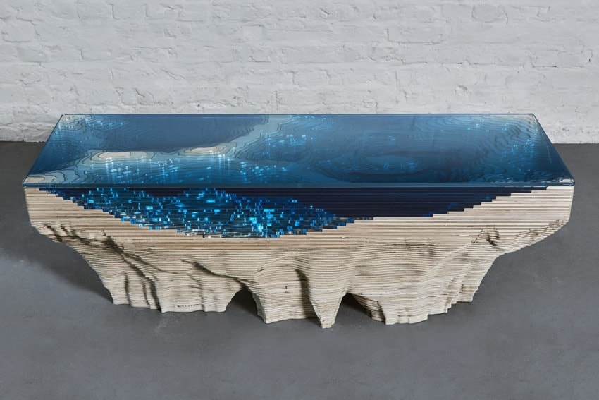 Sculptural coffee table with layered topographic design and blue glass resembling ocean water, set on a gray floor.