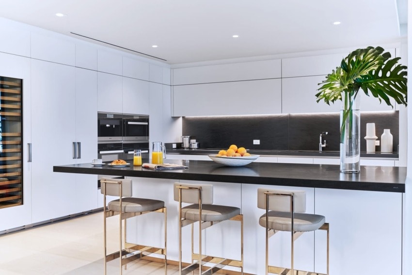 Modern kitchen with white cabinets, black countertop, bar stools, fruit bowl, and sleek appliances.