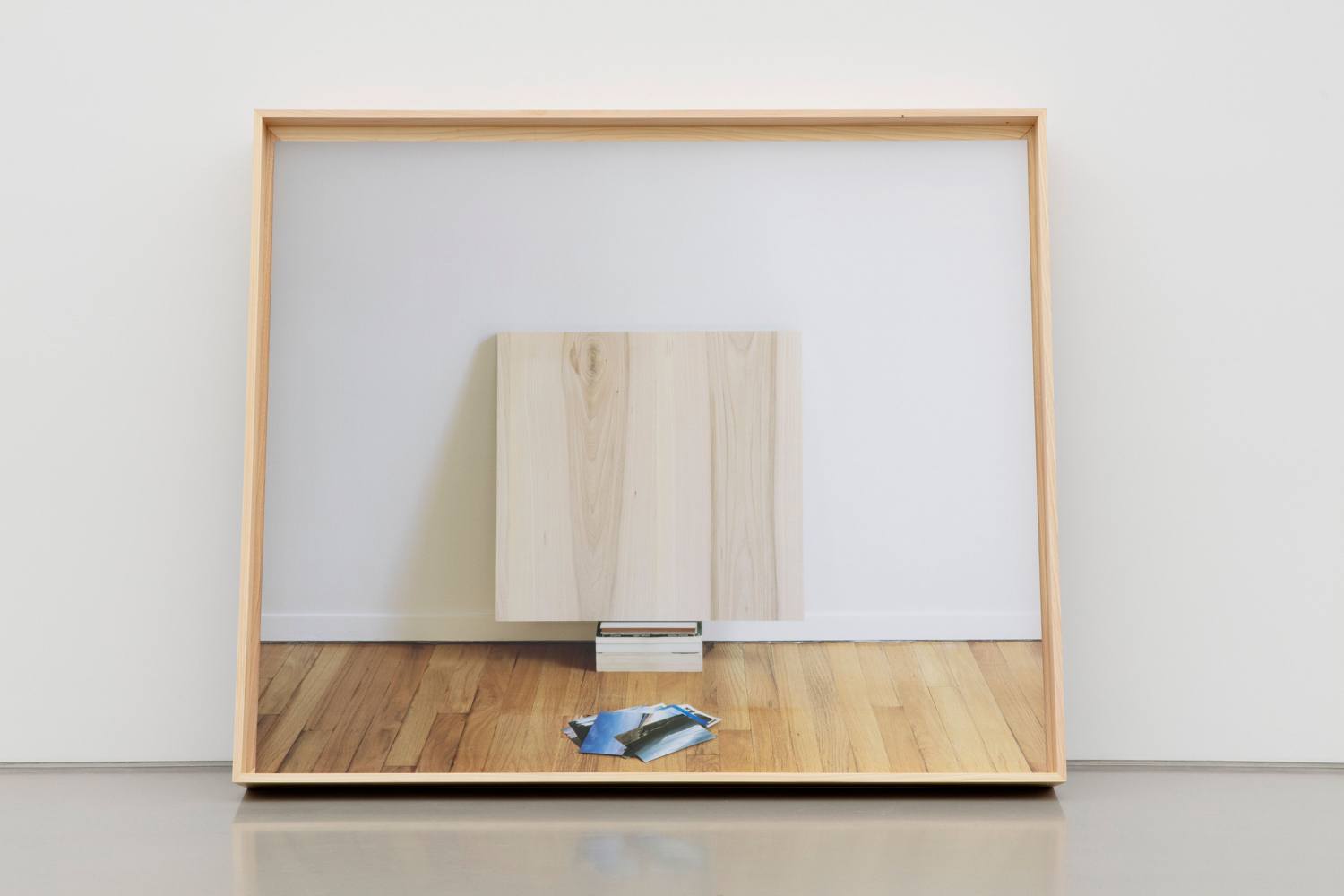 Framed mirror reflecting a wooden block on a wooden floor against a white wall.