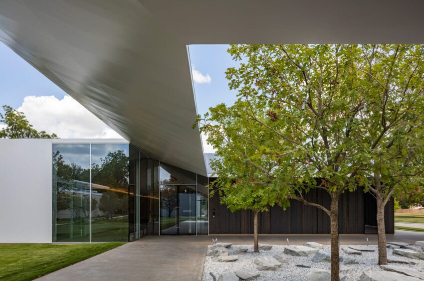 Modern building exterior with glass walls, angular roof, and landscaped area with trees and rocks in the foreground.