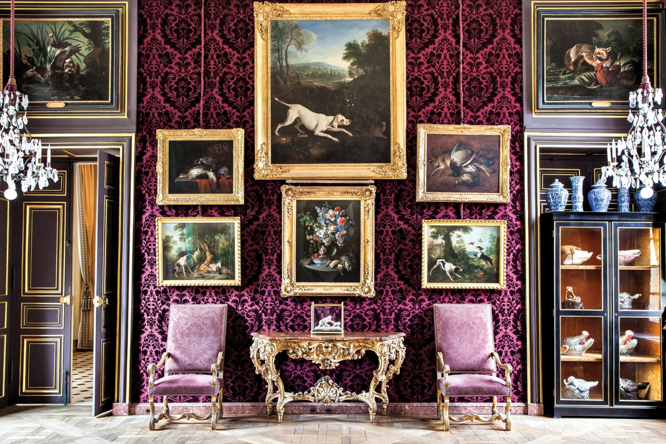 Luxurious room with ornate purple walls, vintage paintings in gold frames, and elegant furniture, including two armchairs.
