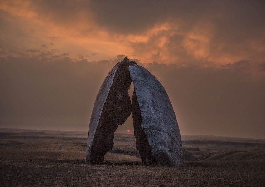 Sculpture resembling a broken egg under a dramatic sunset sky in a vast open landscape.