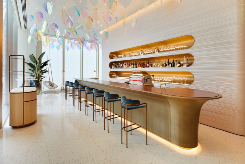 Modern bar interior with a long counter, blue stools, and backlit shelves displaying bottles, under vibrant hanging decorations.