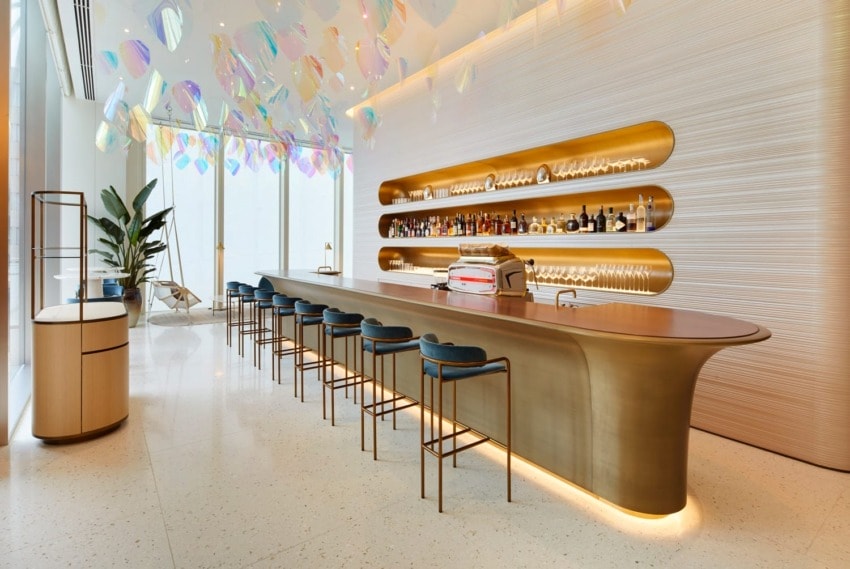 Modern bar interior with a long counter, blue stools, and backlit shelves displaying bottles, under vibrant hanging decorations.
