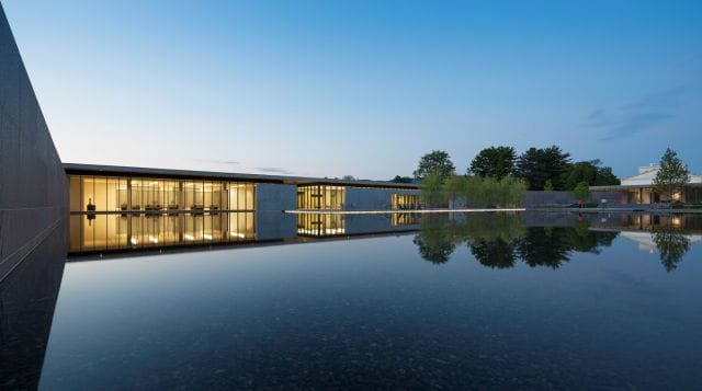 Modern building with glass walls reflecting in a tranquil pond at dusk, surrounded by trees and a clear sky.