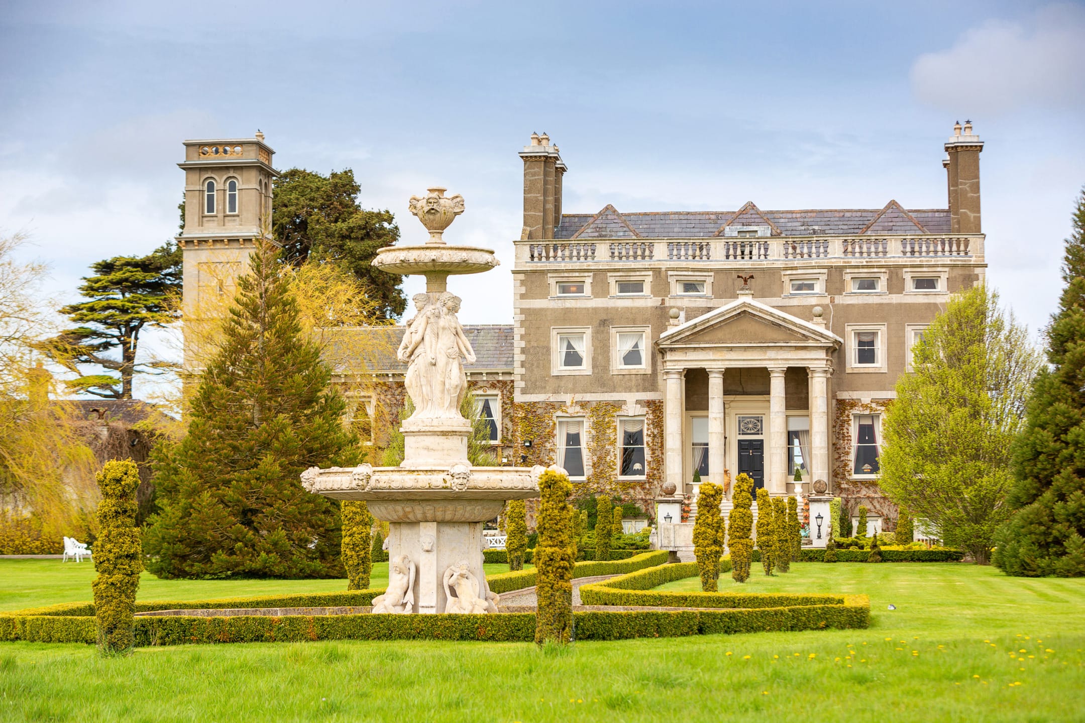 Stately manor with grand entrance, lush gardens, and ornate fountain in foreground on a bright day.