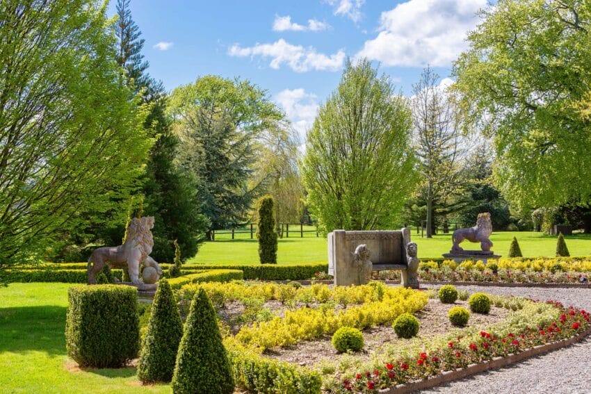 Beautiful garden with colorful flowers, stone lion statues, trimmed hedges, and lush green trees under a sunny blue sky.
