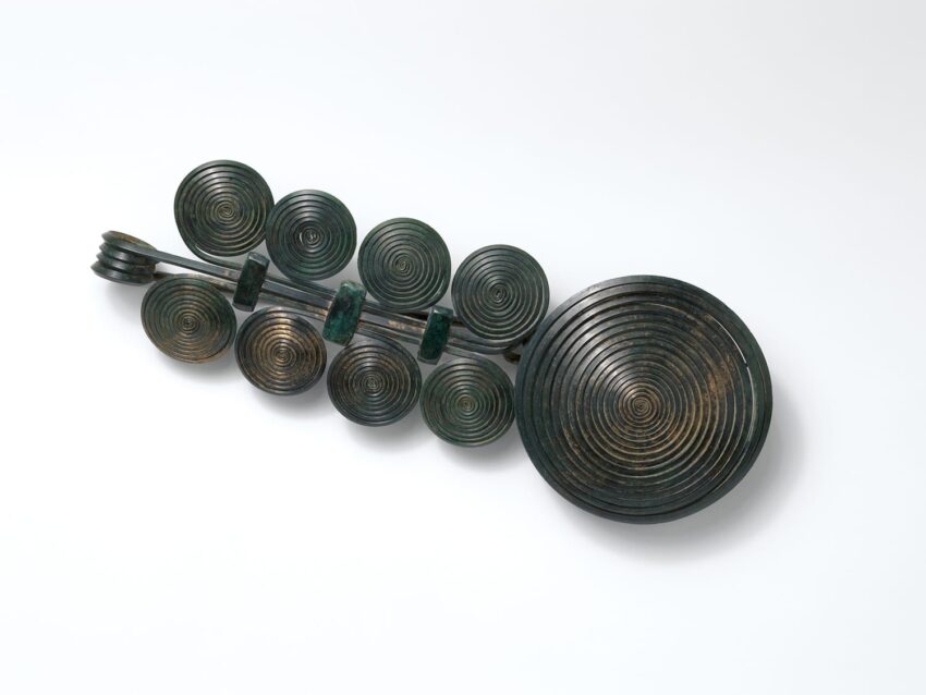Ancient bronze spiral brooch with multiple circular designs arranged symmetrically on a light background.