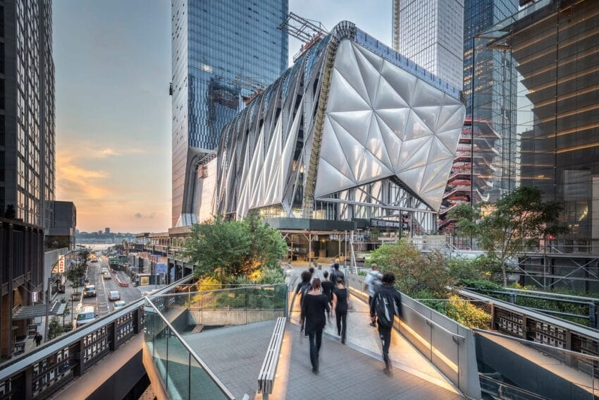Modern architectural building with triangular panels, surrounded by skyscrapers, viewed from a pedestrian walkway at sunset.