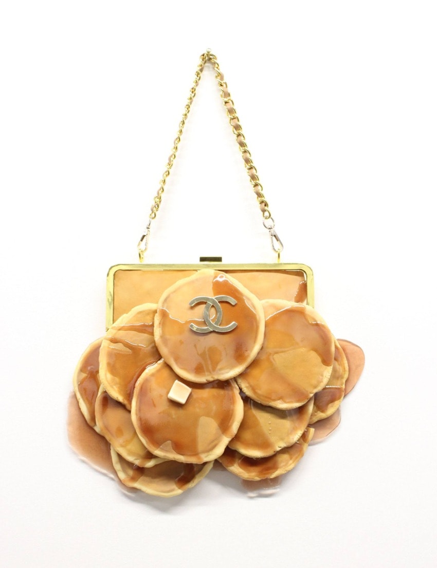 Fashion handbag with pancake and syrup design, gold chain strap, and decorative emblem on a plain background.
