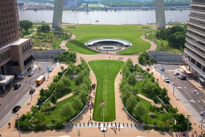 Aerial view of Gateway Arch National Park with people walking, surrounded by lush green spaces and the Mississippi River in the background.