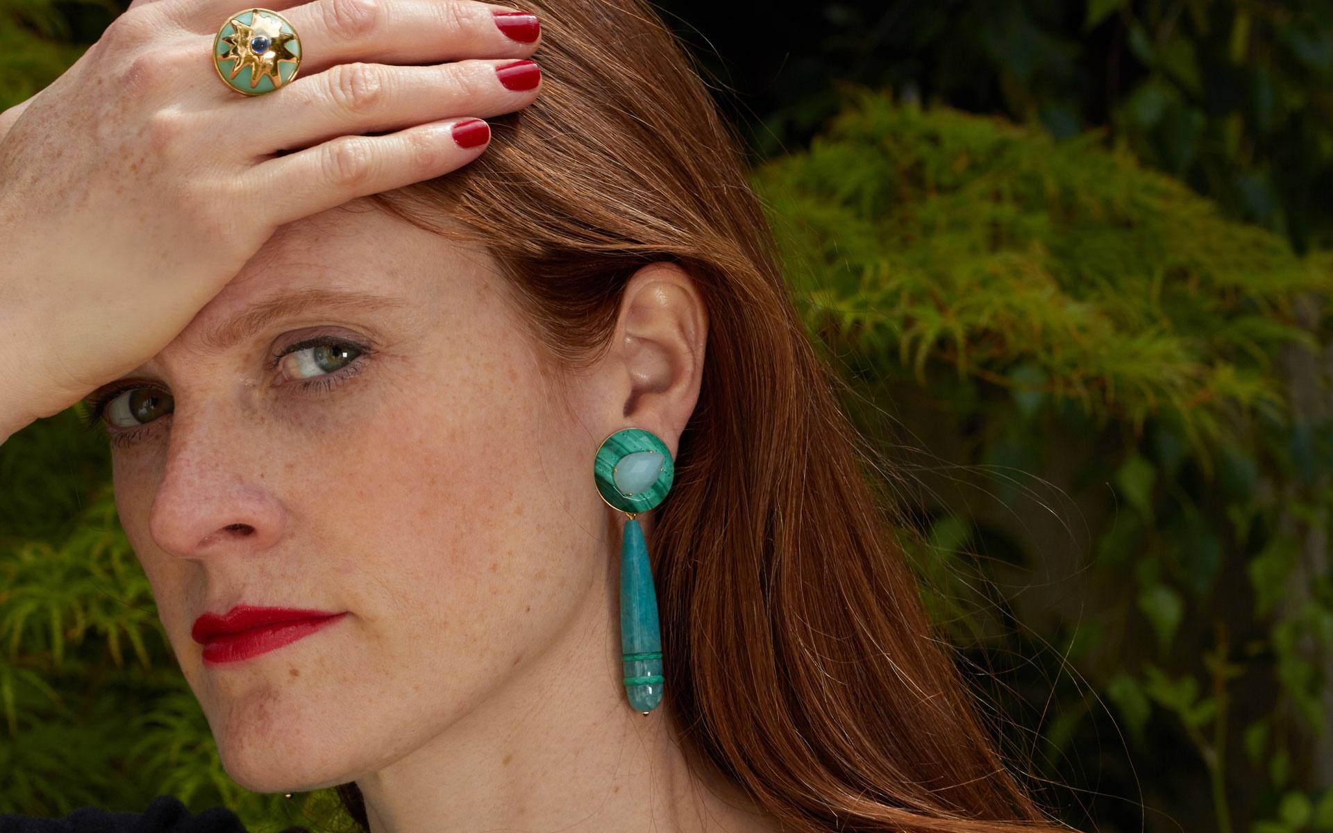 Woman with red hair wearing green earrings and a ring poses with hand on forehead against a leafy background.