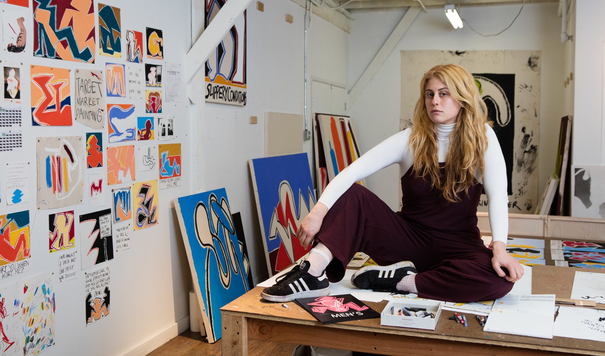 Artist sitting on a table in a studio surrounded by colorful abstract paintings and sketches on the walls.