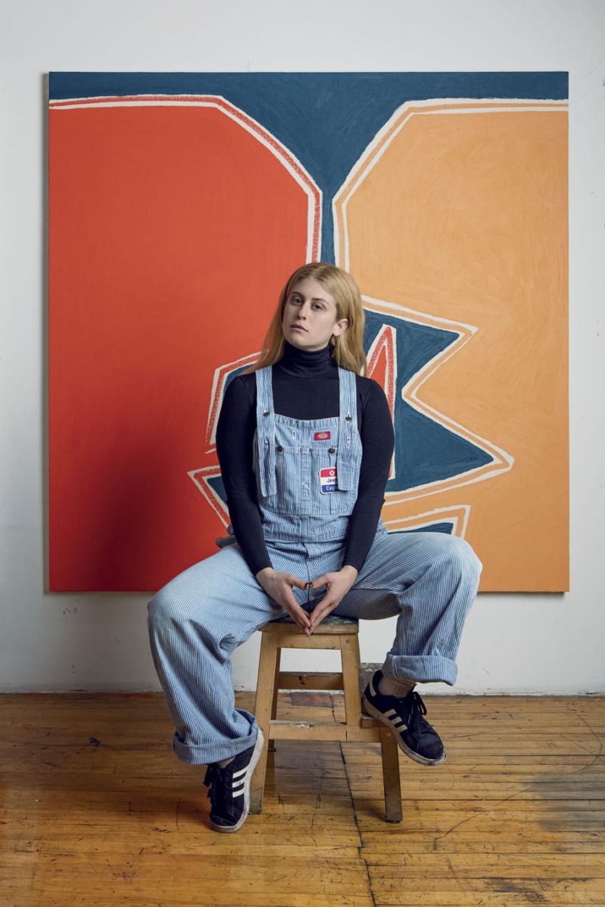 Person in denim overalls sitting on a stool in front of an abstract painting in an art studio.