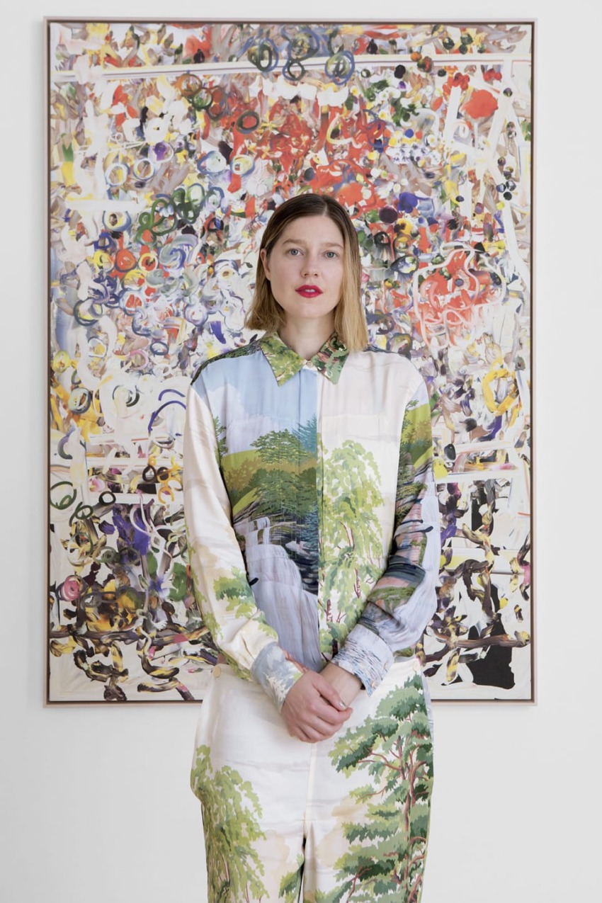 Person in artistic outfit stands in front of colorful abstract painting in a gallery setting.