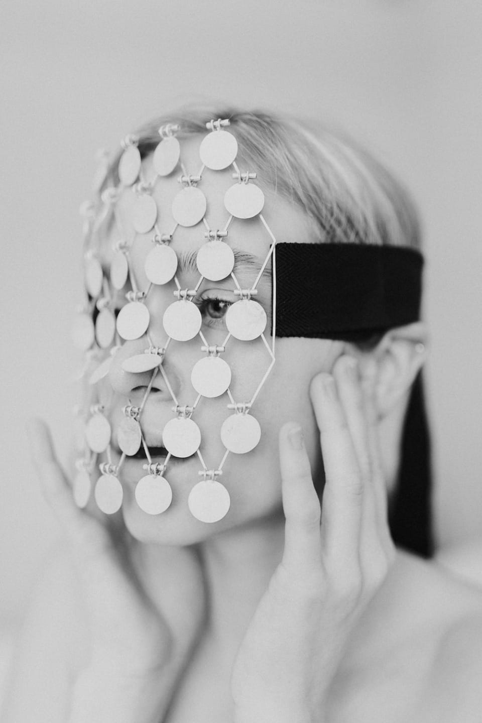 Person wearing a unique face mask made of circular discs, holding it with both hands, black and white photo.