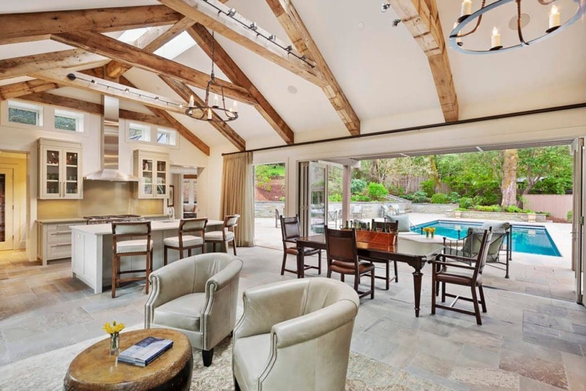 Spacious open-concept room with kitchen, dining area, rustic beams, and view of outdoor pool and garden through glass doors.
