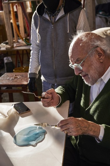 Elderly man creating art with a palette knife on table, while another person stands in the background in a workshop setting.