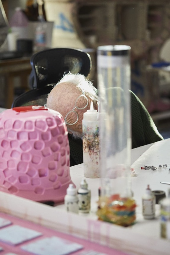 Elderly artist working at cluttered table in studio, surrounded by art supplies and a pink sculpture.