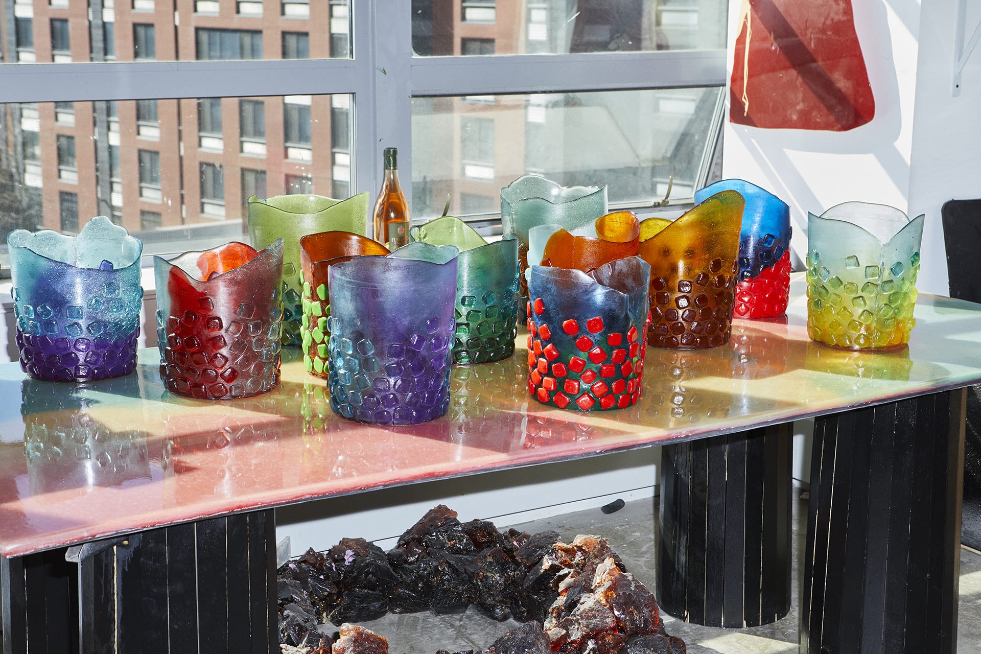 Colorful glass vases on a table in a sunlit room with large windows and a cityscape background.