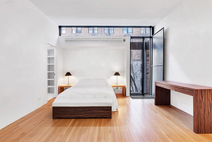 Minimalist bedroom with wooden floor, white walls, simple bed, two lamps, and large window showing urban view.
