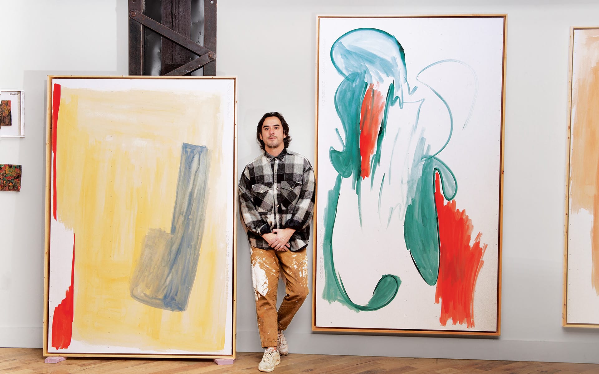 Artist standing next to two abstract paintings, featuring bold colors and expressive brushstrokes, in a gallery setting.