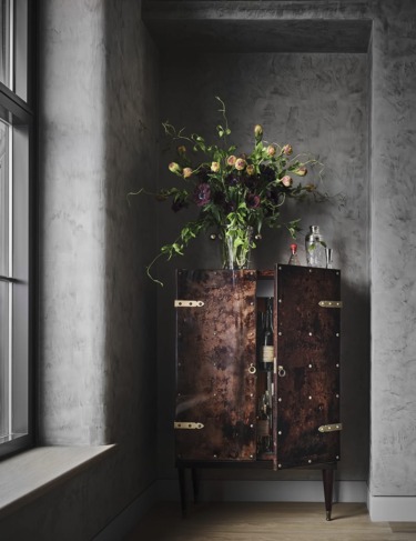 Brown cabinet with open doors revealing wine bottles and glasses, topped with a vase of flowers, in a room with textured walls.
