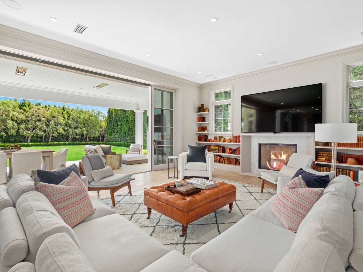Spacious living room with white sofas, a fireplace, large TV, and open doors leading to a patio with outdoor seating and greenery.