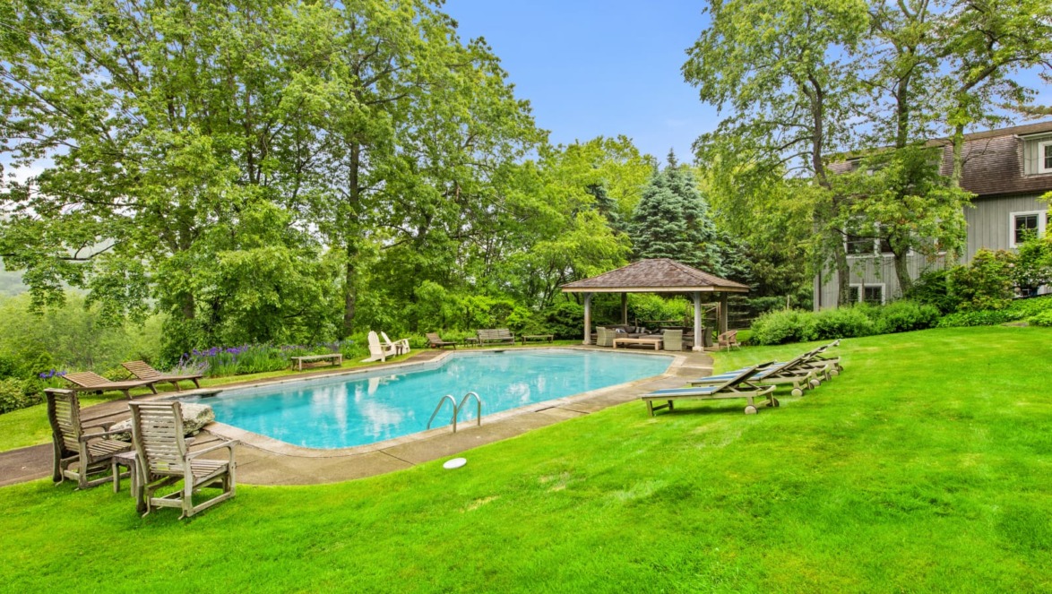 Backyard with a clear swimming pool, surrounded by lush green grass, lounge chairs, and trees under a blue sky.