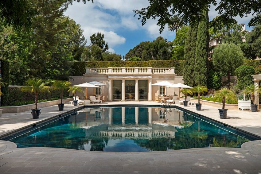 Luxurious outdoor pool area with elegant villa, shaded seating, umbrellas, and lush greenery surrounding the scene.