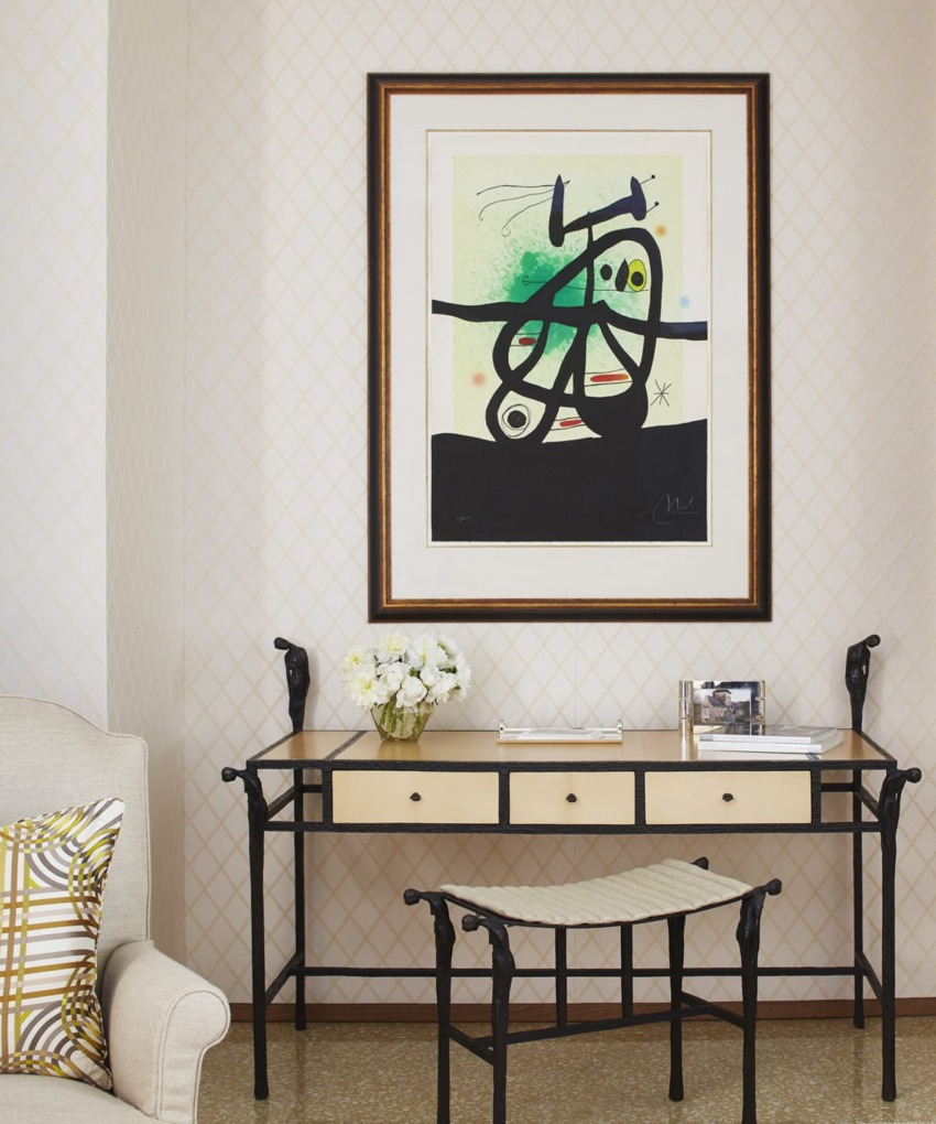 Framed abstract artwork on a wall above a desk with a flower vase, next to a cushioned chair with a patterned pillow.