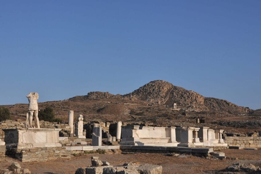 Ancient ruins with statues set against a rocky hill under a clear blue sky.